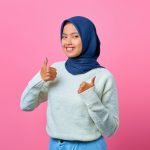 Smiling young woman in hijab and white sweater giving thumbs up on a pink background in a studio setting.