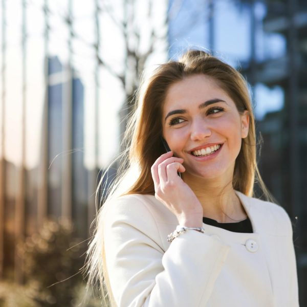 pexels-photo-3754676-3754676 Happy woman in white coat chatting on mobile phone in an urban setting with sunlight.