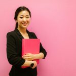 Smiling Asian woman holding a pink binder against a pink background, conveying positivity.